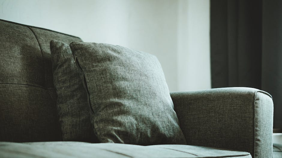 Close-up image of a modern grey upholstered sofa with a matching cushion placed on it, situated in a well-lit living room with natural light streaming in from the right side. The fabric of the sofa ap